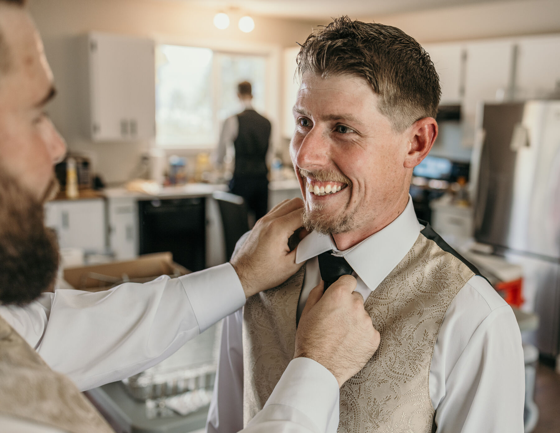 Groom laughing as a groomsman straightens his tie in a kitchen