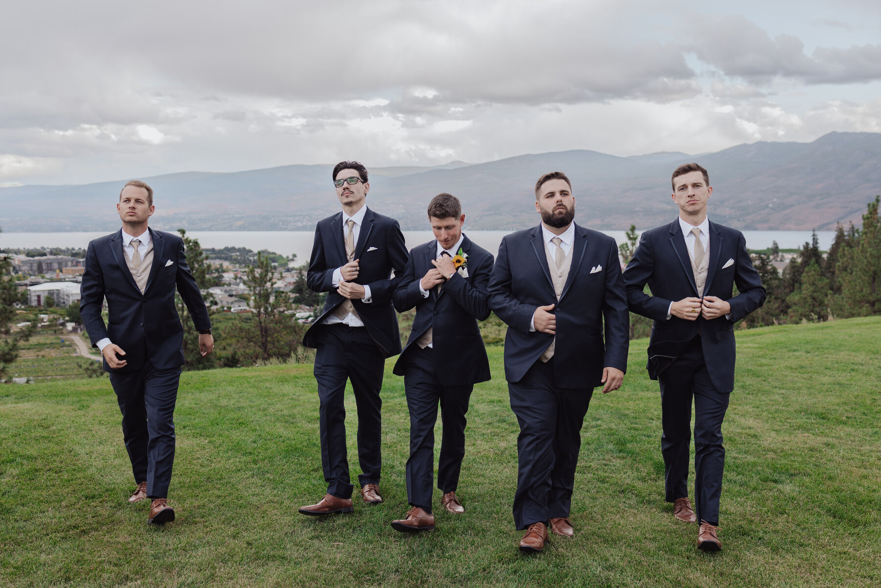Groomsmen in navy suits walking toward the camera across a grassy hillside