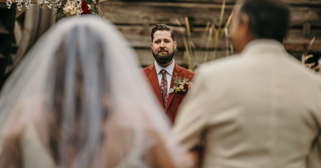 Groom in a burgundy suit watching his bride walk down the aisle