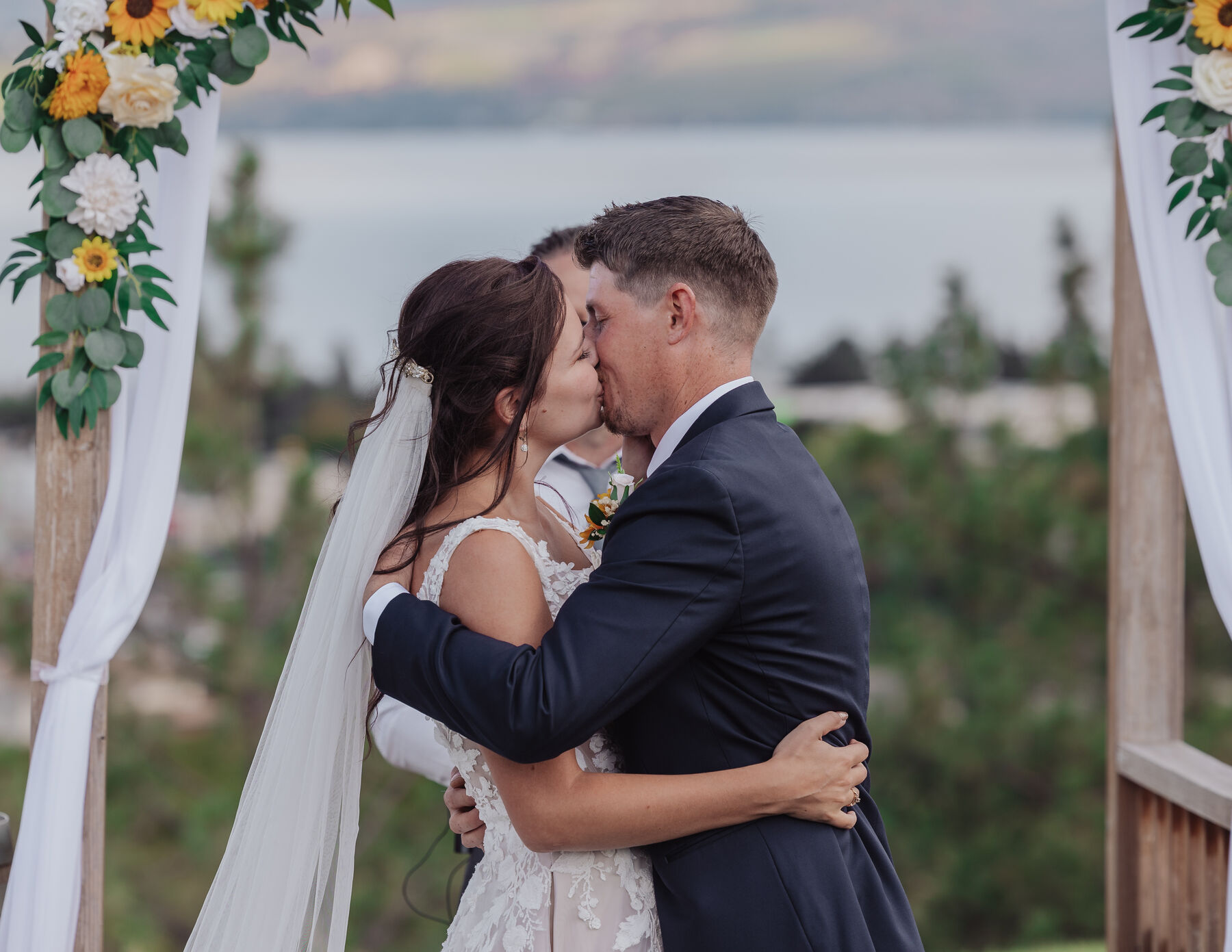 Bride and groom kissing under a sunflower-decorated arch overlooking the lake