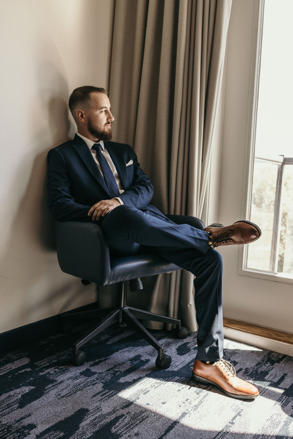 Groom in a navy suit relaxing in a chair by a sunlit window