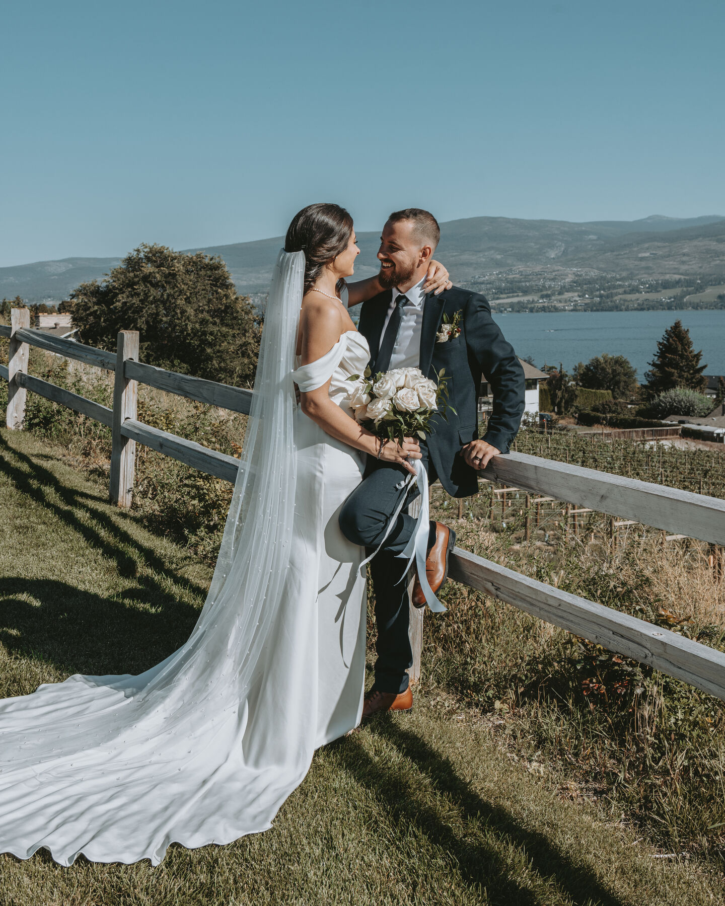 Bride and groom leaning on a fence above the lake, her long veil flowing