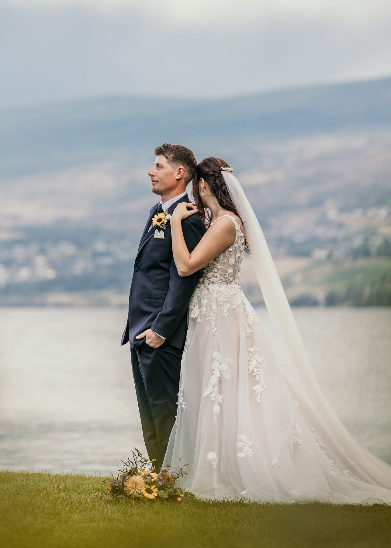Bride and groom standing close beside the lake, her veil blowing, a bouquet at their feet